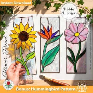 May include: Three rectangular stained glass panels featuring floral designs: a yellow sunflower, an orange and purple bird of paradise, and a pink flower. Each panel has a dark metal frame and textured glass background, displayed on a white sheet.