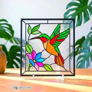 May include: A stained glass panel featuring a colorful hummingbird with green, red, and orange plumage, hovering near a vibrant pink and blue flower. The artwork is framed in black and includes green leaves. The text "By: QGCrafts" is at the bottom.