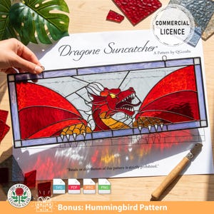 Op de afbeelding: Een gebrandschilderde glazen drakenvanger met een rood drakenontwerp. De draak heeft open vleugels en staat tegen een lichte achtergrond. De tekst "Dragone Suncatcher" en "Commercial Licence" zijn zichtbaar. Inclusief een bonus kolibrie patroon.