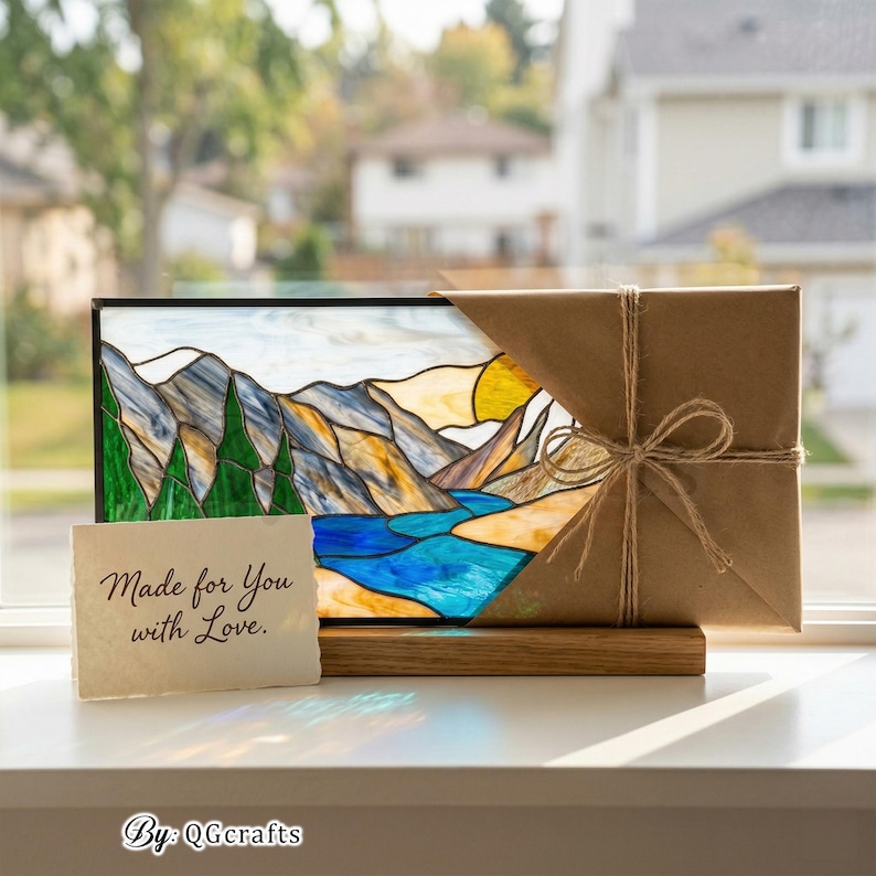 Peut inclure: Vitrail repr&eacute;sentant un paysage de montagnes, une rivi&egrave;re et un soleil. L'&oelig;uvre est pr&eacute;sent&eacute;e sur un socle en bois, avec un cadeau envelopp&eacute; dans du papier brun et ficel&eacute; avec de la ficelle. Une petite carte indique "Made for You with Love."