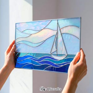 以下が含まれることがあります： 海に浮かぶヨットを描いたステンドグラスパネル。青と白の色合いで、中央にヨットが描かれています。「By: QGcrafts」の文字が隅にあります。