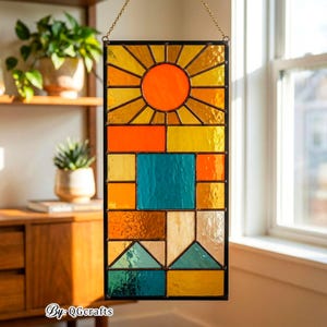 May include: A rectangular stained glass panel with a sunburst design in shades of yellow and orange, a turquoise square, and geometric shapes in various colors. The panel is framed in black and hangs from a gold chain. The text "By: QGcrafts" is at the bottom.