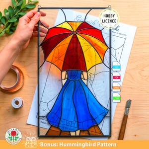 Puede incluir: Vidriera que representa a una mujer con un vestido azul sosteniendo un paraguas colorido. El paraguas tiene secciones rojas, naranjas y amarillas. La obra está enmarcada en negro e incluye el texto "Bonus: Hummingbird Pattern".