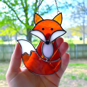 Puede incluir: Atrapasoles de vidrio con forma de zorro en tonos naranja, blanco y negro. El zorro tiene una expresión alegre y está suspendido por una cadena. Este atrapasoles está diseñado para capturar la luz y añadir un toque decorativo.
