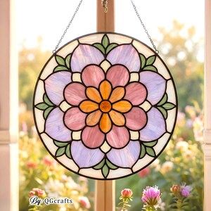 May include: A round stained glass window with a floral design. The window features a central orange flower surrounded by pink and purple petals, green leaves, and a white border. The text "By: QGcrafts" is visible at the bottom.