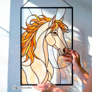 May include: Stained glass panel featuring a horse head with a flowing orange and yellow mane. The horse is depicted in shades of cream and tan, outlined in black. The panel is framed in black and suspended by a chain. Text in the lower left corner reads "By: QGcrafts."