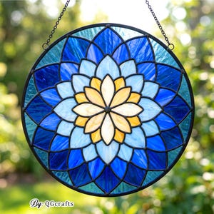 May include: A round stained glass window with a floral design in shades of blue, light blue, yellow, and white. The glass is framed with a black metal border and hangs from a chain. The text "By: QGCrafts" is in the bottom left corner.