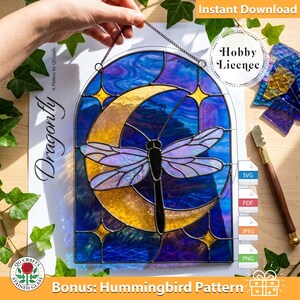 May include: A stained glass suncatcher featuring a dragonfly centered over a yellow crescent moon against a deep blue background with small yellow stars. The piece is arched at the top and includes a metal chain for hanging. The design is displayed on a white paper pattern sheet labeled Dragonfly with a Hobby Licence icon.