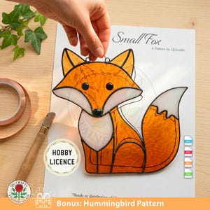 May include: A stained glass fox with orange and white coloring, black eyes, and a black nose. The fox is hanging in front of a paper with the text "Small Fox" and "A Pattern by QGcrafts". The image also includes the text "Bonus: Hummingbird Pattern".
