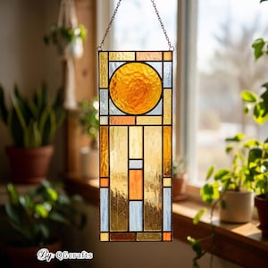 May include: A rectangular stained glass panel with a geometric design. The panel features a large, circular sun in the center, surrounded by rectangular and square shapes in shades of yellow, orange, and blue. The panel is suspended by a chain.