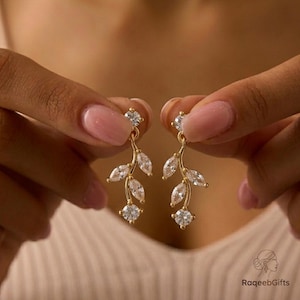May include: Gold earrings with a leaf design, adorned with clear, sparkling gemstones. The earrings have a delicate, elegant appearance, with a combination of round and marquise-cut stones. The design is reminiscent of a natural, botanical theme.