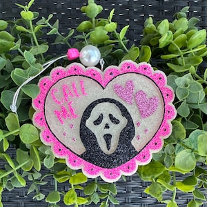 May include: A heart-shaped air freshener with a pink scalloped border and glitter accents. The design features the Ghostface mask from the Scream movies, the text "CALL ME", and two glitter hearts. A white pearl and pink bead are attached.