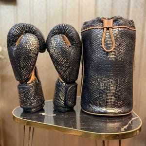 以下が含まれることがあります： 黒と金のボクシンググローブと、お揃いの円筒形のバッグ。グローブとバッグはヘビ革の質感です。バッグには巾着の留め具と「PYTHOS ATHLETICS」の文字が入っています。セットは金色の縁取りのテーブルに飾られています。