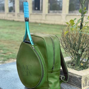 Pode incluir: Mochila de ténis em couro verde com uma raquete de ténis verde a condizer. A raquete tem uma pega azul. A mochila tem um bolso com zíper para a raquete e um compartimento principal com zíper. A mochila tem as letras "MC ON" em relevo na lateral.