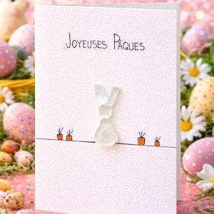 May include: White Easter greeting card with the words "Joyeuses Pâques" and a sea glass bunny design. The card features a textured surface, a black line with carrot illustrations, and is surrounded by Easter eggs and flowers.
