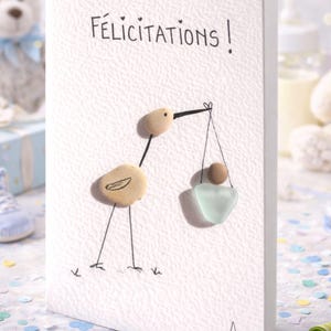 Könnte beinhalten: Eine weiße Grußkarte mit dem Wort "Félicitations!" und einer Illustration eines Storchs aus Kieselsteinen. Der Storch trägt ein Baby in einer Tragetasche aus Meeresglas. Die Karte hat eine strukturierte Oberfläche.
