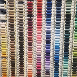 Può includere: Un'esposizione a parete di rocchetti di filo in una vasta gamma di colori. I fili sono disposti in colonne verticali, mostrando uno spettro che va dal bianco al nero, passando poi per varie tonalità di rosso, rosa, blu, verde e giallo.