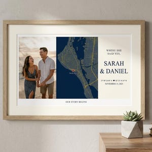 May include: A framed print featuring a photograph of a couple and a map. The map highlights a coastal area, marked with a heart. Text includes "Where she said yes," names, and a date. The frame is light brown.