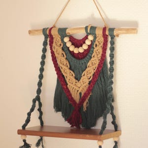 Può includere: Una mensola da parete in macramè con un ripiano in legno e un design decorativo intrecciato. Il macramè presenta una combinazione di colori verde acqua, bordeaux e beige, con perline e nappe in legno. La mensola è sospesa da un cordoncino in fibra naturale.