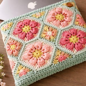 Może przedstawiać: Szydełkowy pokrowiec na laptopa z kwiatowym wzorem w odcieniach różu, żółci i brzoskwini na jasnym zielonym tle. Pokrowiec ma drewniane zapięcie na guzik i jest przeznaczony do laptopa.