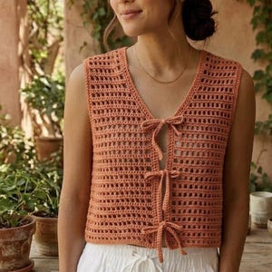 Può includere: Un top all'uncinetto senza maniche color pesca con scollo a V e chiusure con fiocco sul davanti. Il design a trama aperta del top è visibile. Il top è abbinato a pantaloni bianchi. Lo sfondo comprende piante in vaso e un muro di pietra.