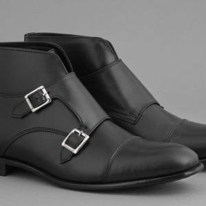 May include: A pair of black leather ankle boots with silver-colored buckles. The boots have a cap toe design and a smooth, polished finish. The buckles are rectangular and secure the straps across the top of the foot.