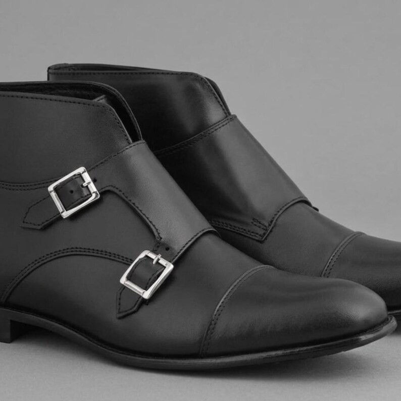 May include: A pair of black leather ankle boots with silver-colored buckles. The boots have a cap toe design and a smooth, polished finish. The buckles are rectangular and secure the straps across the top of the foot.
