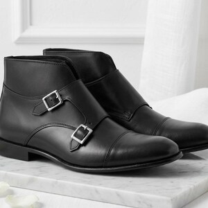 May include: A pair of black leather ankle boots with silver-tone buckles. The boots have a cap toe design and are displayed on a marble surface. The footwear is a classic style.