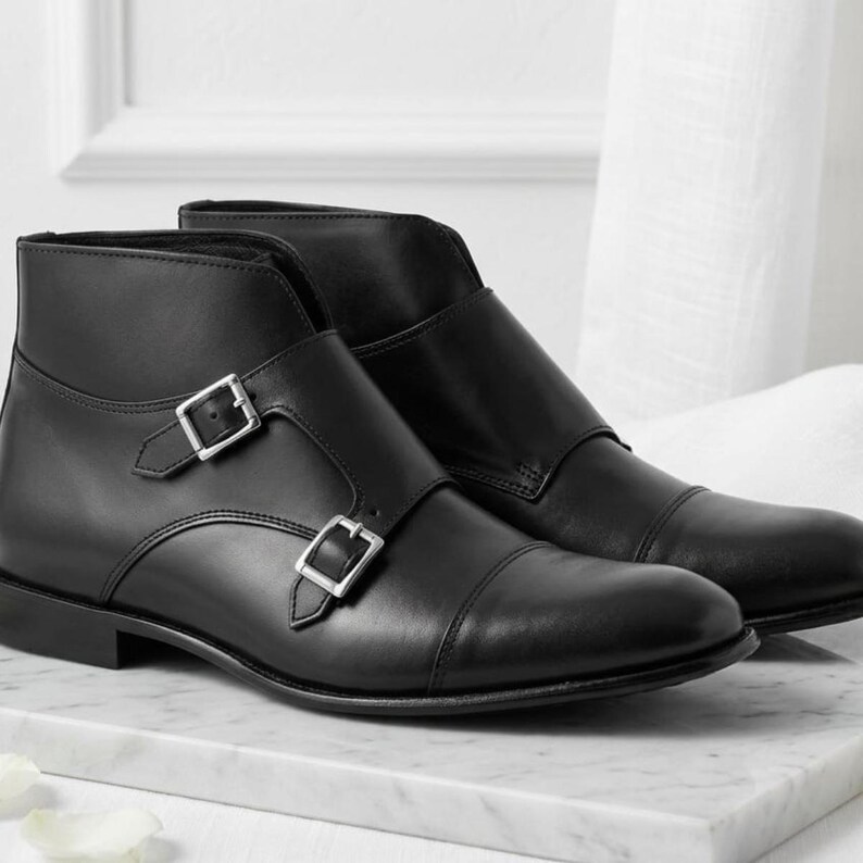 May include: A pair of black leather ankle boots with silver-tone buckles. The boots have a cap toe design and are displayed on a marble surface. The footwear is a classic style.