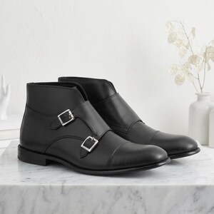 May include: A pair of black leather ankle boots with double buckle closures. The boots have a cap toe design and are displayed on a marble surface. The footwear is a classic style.