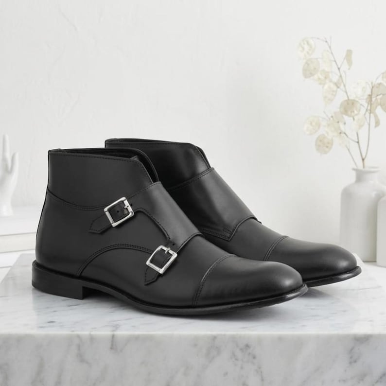 May include: A pair of black leather ankle boots with double buckle closures. The boots have a cap toe design and are displayed on a marble surface. The footwear is a classic style.