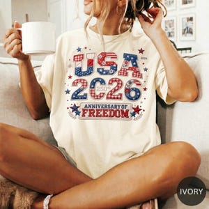 May include: Ivory-colored t-shirt with a patriotic design. The shirt features the text "USA 2026" in red, white, and blue, celebrating the 250th Anniversary of Freedom. The design includes stars and a banner.