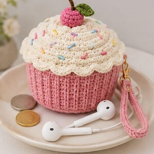 May include: A crocheted cupcake-shaped pouch featuring a pink ribbed base and a cream-colored top decorated with colorful sprinkles. A small pink cherry with a green leaf sits on top. A pink crocheted wrist strap with a gold clasp is attached.
