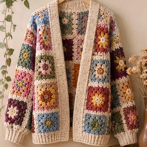 Könnte beinhalten: Eine langärmelige Strickjacke aus mehrfarbigen Häkel-Granny-Squares mit Blumenmustern in Gelb-, Blau-, Rosa-, Grün- und Burgundertönen. Das Kleidungsstück hat einen cremefarbenen Rippsaum an der Vorderöffnung und an den Ärmelbündchen.