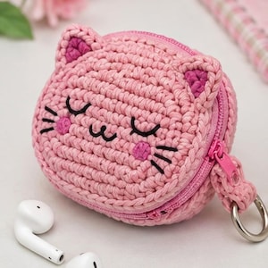 May include: A round, pink crocheted case shaped like a cat face with two ears, embroidered black eyes, whiskers, a mouth, and pink cheeks. The case features a pink zipper closure along the side and a silver metal ring attached to the side for hanging.