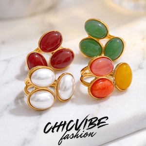Peut inclure: Anneaux dorés ornés de pierres ovales rouges, vertes, oranges, jaunes et blanches. Les bagues présentent un design à double bande et sont disposées sur une surface blanche. Le texte "CHICVIBE fashion" est visible.