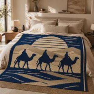 May include: A woven throw blanket featuring a dark blue and beige desert scene. The design displays three silhouettes of people riding camels across sand dunes under a large, striped circular sun. The blanket is spread flat over a bed.