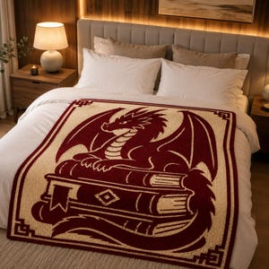 Puede incluir: Una manta rectangular con un dragón de color rojo oscuro posado sobre una pila de libros sobre un fondo color crema. El diseño incluye un borde decorativo con detalles geométricos en las esquinas, todo con textura de punto.