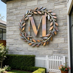 Puede incluir: Una gran letra "M" metálica está montada en una pared de piedra, rodeada por una corona decorativa de hojas y bayas. El metal tiene un acabado degradado de tonos bronce, oro y plata. Una valla blanca y un jardín verde están en primer plano.