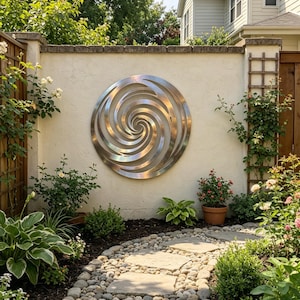 Op de afbeelding: Een groot, cirkelvormig metalen wandkunstwerk met een spiraalontwerp is bevestigd aan een crèmekleurige muur. Het kunstwerk is omgeven door weelderig groen, waaronder struiken, bloemen en een stenen pad. De tuin is goed verlicht met natuurlijk zonlicht.