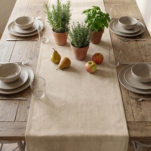 Puede incluir: Una mesa de comedor rústica con un camino de mesa de lino beige. La mesa está puesta con platos, cuencos y vasos beige. Hierbas en macetas y fruta fresca, como peras y manzanas, añaden un toque natural a la decoración.