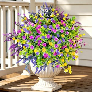 May include: A round arrangement of artificial flowers in a white pedestal planter. The bouquet features a dense mix of vibrant yellow, bright purple, and magenta blossoms interspersed with green foliage and tall, slender purple lavender-like stems.