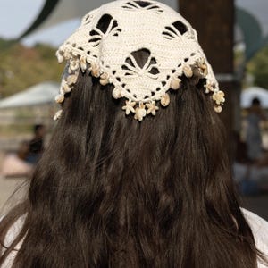 Peut inclure: Un foulard triangulaire en crochet blanc cassé présentant un motif ajouré complexe et une bordure décorative ornée de petits coquillages clairs et d'accents en forme d'étoile suspendus le long du bord inférieur du tissu.
