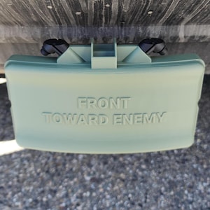 Pode incluir: Um objeto retangular verde claro com as palavras "FRONT TOWARD ENEMY" em relevo na frente. O objeto tem duas protuberâncias pretas e arredondadas no topo. O objeto está sobre uma superfície de pequenas pedras cinzentas.