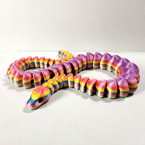 May include: A colorful, articulated snake toy in a rainbow gradient of red, orange, yellow, green, blue, and purple. The segmented body allows for flexible posing. The snake's head is detailed with black eyes.