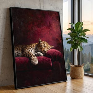 May include: A framed wall art print featuring a leopard with a spotted coat sleeping on a deep red velvet sofa. The background is a dark, textured burgundy wall. The artwork is enclosed in a thin black frame and rests against a concrete wall.
