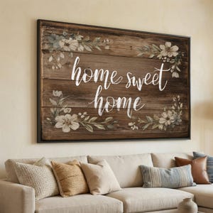 May include: A rectangular wooden sign with the words "home sweet home" in white script. The sign is framed in black and decorated with floral accents in shades of white and green. The sign is hanging above a beige sofa with decorative pillows.