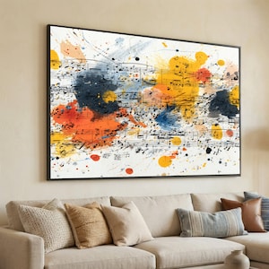 Op de afbeelding: Een ingelijst canvas kunstwerk met muzieknoten en kleurrijke verfspatten in geel, oranje, blauw en zwart. Het kunstwerk hangt boven een beige bank met diverse kussens.