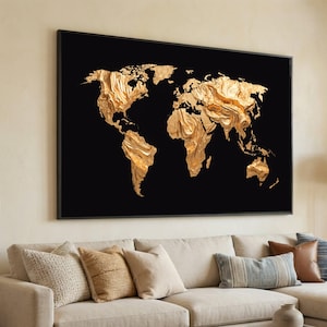 May include: A large framed wall art piece featuring a gold-colored world map against a black background. The map has a textured, three-dimensional appearance. The artwork is displayed above a beige sofa with decorative pillows.