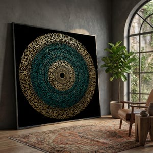 May include: Large framed artwork featuring intricate gold and teal calligraphy on a black background. The circular design is centered, with the calligraphy spiraling outwards. The artwork is displayed in a room with a rug, chair, and a potted plant.